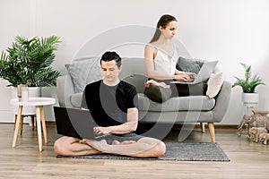 Young couple in sportswear, sitting in lotus pose, man on the floor and woman on gray sofa, listening yoga practices