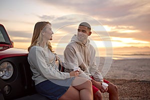 Young Couple Standing Chatting By Car With Hot Drink At Beach Watching Sunrise Together photo