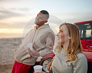 Young Couple Standing Chatting By Car With Hot Drink At Beach Watching Sunrise Together photo