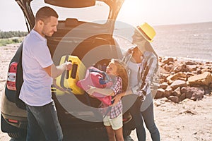 Young couple standing near the opened car boot with suitcases and bags. Dad, mom and daughter are traveling by the sea