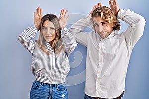 Young couple standing over blue background doing bunny ears gesture with hands palms looking cynical and skeptical