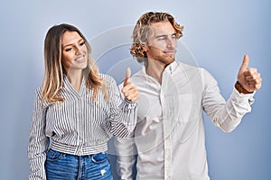 Young couple standing over blue background looking proud, smiling doing thumbs up gesture to the side