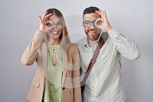 Young couple standing over white background doing ok gesture with hand smiling, eye looking through fingers with happy face
