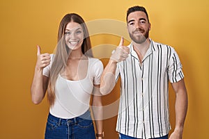 Young couple standing over yellow background doing happy thumbs up gesture with hand