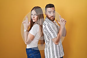 Young couple standing over yellow background holding symbolic gun with hand gesture, playing killing shooting weapons, angry face