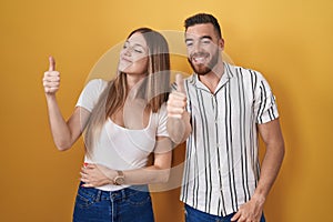 Young couple standing over yellow background looking proud, smiling doing thumbs up gesture to the side