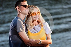 young couple in sunglasses hugging on river beach in evening