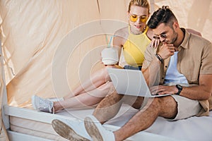 young couple in sunglasses using laptop while sitting