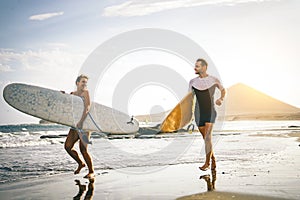 Young couple of surfers running with surfboards on the beach at sunset - Happy lovers going to surf together