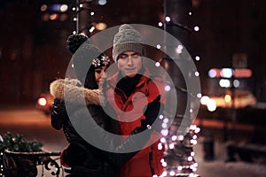 Young couple in sweaters and caps embrace on the winter street. In love in a Christmas garland. St. Valentine`s Day