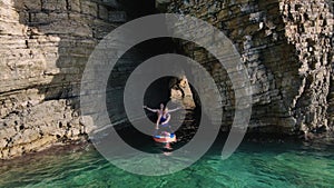 Young couple swims on SUP board through crevice between cliffs in sea. Woman is rowing with oar, man is chilling with