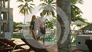 Young couple in swimsuits walking along swimming pool of luxury resort hotel on ocean shore. Cheerful girl and guy
