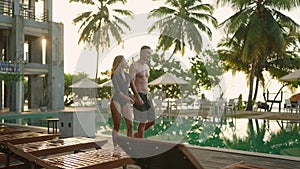 Young couple in swimsuits walking along swimming pool of luxury resort hotel on ocean shore. Cheerful girl and guy