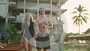 Young couple in swimsuits walking along swimming pool of luxury resort hotel on ocean shore. Cheerful girl and guy