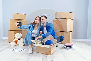 Young couple taking selfies in their new home while sitting among cardboard boxes. Happy attractive young couple is