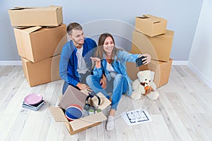 Young couple taking selfies in their new home while sitting among cardboard boxes. Happy attractive young couple is