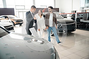 Young couple talking to sales person in car showroom choosing automobile