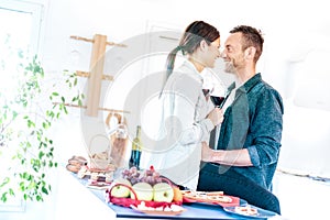 Young couple toasting red wine and almost kissing at home kitchen - Happy millenial lovers enjoying tender moment cheering