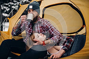 Young couple in tourist tent during winter hike. Guy drinks hot tea from metal mug, girl lies.