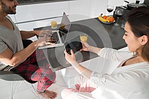 Young Couple Using Cell Smart Phone And Laptop Computer , Young Woman Man Morning Sitting At Kitchen Table