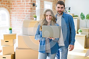 Young couple using computer laptop standing on a room around cardboard boxes, happy for moving to a new apartment