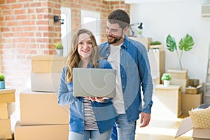 Young couple using computer laptop standing on a room around cardboard boxes, happy for moving to a new apartment