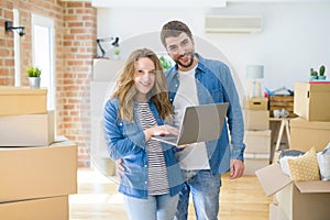 Young couple using computer laptop standing on a room around cardboard boxes, happy for moving to a new apartment