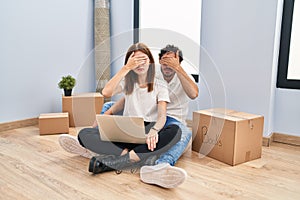 Young couple using laptop at new home covering eyes with hand, looking serious and sad