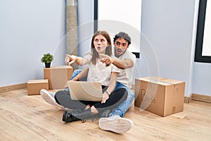 Young couple using laptop at new home pointing with finger surprised ahead, open mouth amazed expression, something on the front