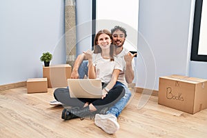 Young couple using laptop at new home pointing to the back behind with hand and thumbs up, smiling confident