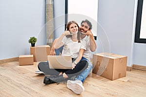 Young couple using laptop at new home smiling doing phone gesture with hand and fingers like talking on the telephone