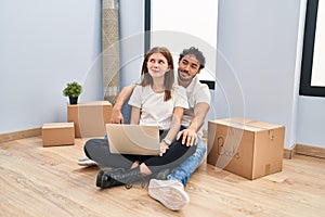 Young couple using laptop at new home smiling looking to the side and staring away thinking