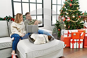 Young couple using laptop sitting by christmas tree checking the time on wrist watch, relaxed and confident