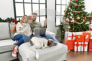 Young couple using laptop sitting by christmas tree cheerful with a smile on face pointing with hand and finger up to the side