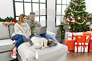 Young couple using laptop sitting by christmas tree with a happy and cool smile on face