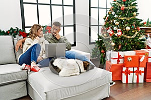 Young couple using laptop sitting by christmas tree looking stressed and nervous with hands on mouth biting nails