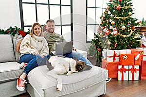 Young couple using laptop sitting by christmas tree winking looking at the camera with sexy expression, cheerful and happy face