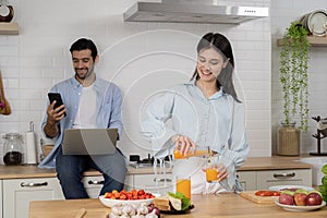 Young couple using laptop and smartphone in the kitchen, smiling man working online at home while his wife cooking with phone apps