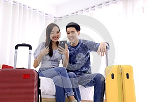 Young couple using mobile phone while sitting together on bed at a hotel room with a suitcase. Couple of tourists searching