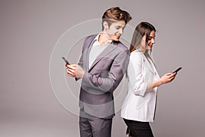 Young couple is using smart phones and smiling while standing back to back on a gray background. Man look at woman.
