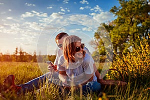 Young couple using their phones and laughing outside. Woman and man relaxing after having picnic at sunset.