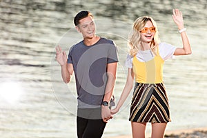 young couple walking on river beach in evening and waving hands