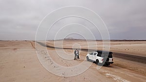 Young couple walks on highway, car at roadside parked in desert. White SUV.