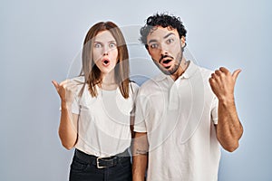 Young couple wearing casual clothes standing together surprised pointing with hand finger to the side, open mouth amazed