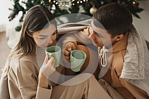 Young couple wearing comfy loungewear drinking tea near Christmas tree