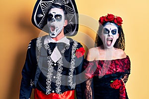 Young couple wearing mexican day of the dead costume over yellow angry and mad screaming frustrated and furious, shouting with