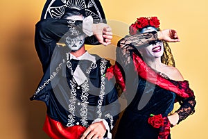 Young couple wearing mexican day of the dead costume over yellow covering eyes with arm smiling cheerful and funny