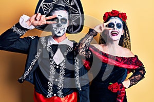 Young couple wearing mexican day of the dead costume over yellow doing peace symbol with fingers over face, smiling cheerful