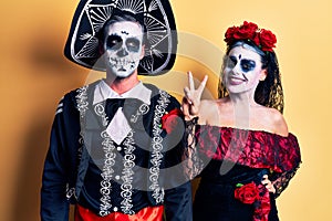 Young couple wearing mexican day of the dead costume over yellow showing and pointing up with fingers number two while smiling