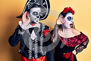 Young couple wearing mexican day of the dead costume over yellow smiling with hand over ear listening an hearing to rumor or